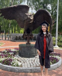 A young woman in a cap and gown standing in front of an owl statue. 