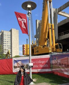 A young woman in a graduation gown standing in front of a building with a College of Public Health banner.