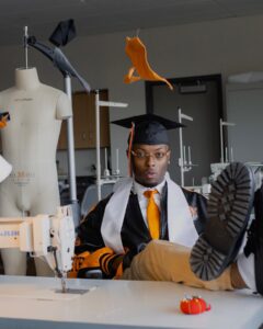 young man in graduation cap sitting at a sewing machine. 