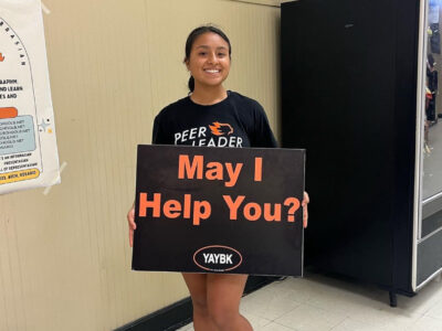 PeerForward Peer Leader Paulina Anaya poses with a sign that reads "May I Help You"