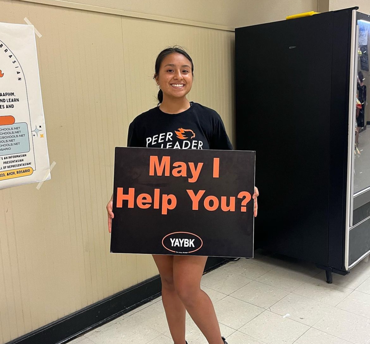 PeerForward Peer Leader Paulina Anaya poses with a sign that reads "May I Help You"